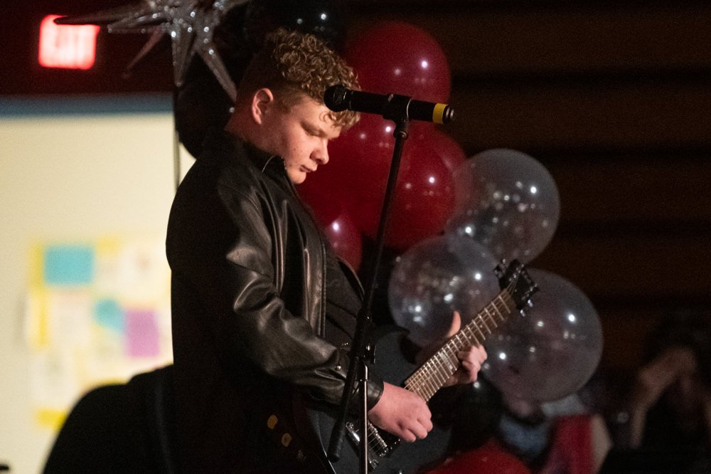 Young guitarist in a black leather jacket playing an electric guitar on stage, with red and silver balloons in the background.