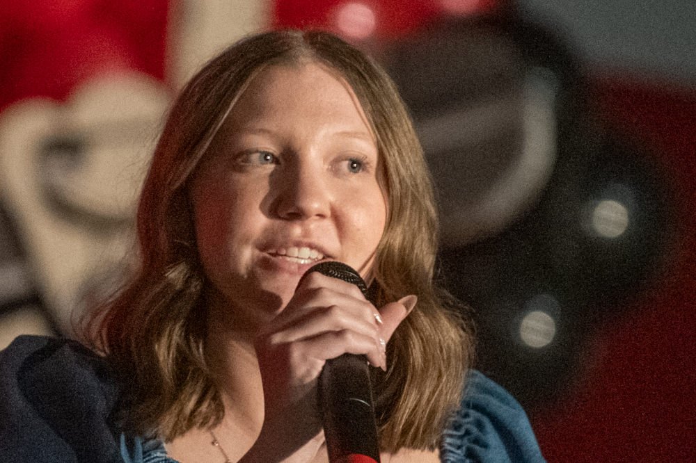 Woman with shoulder-length hair speaking into a handheld microphone on a stage