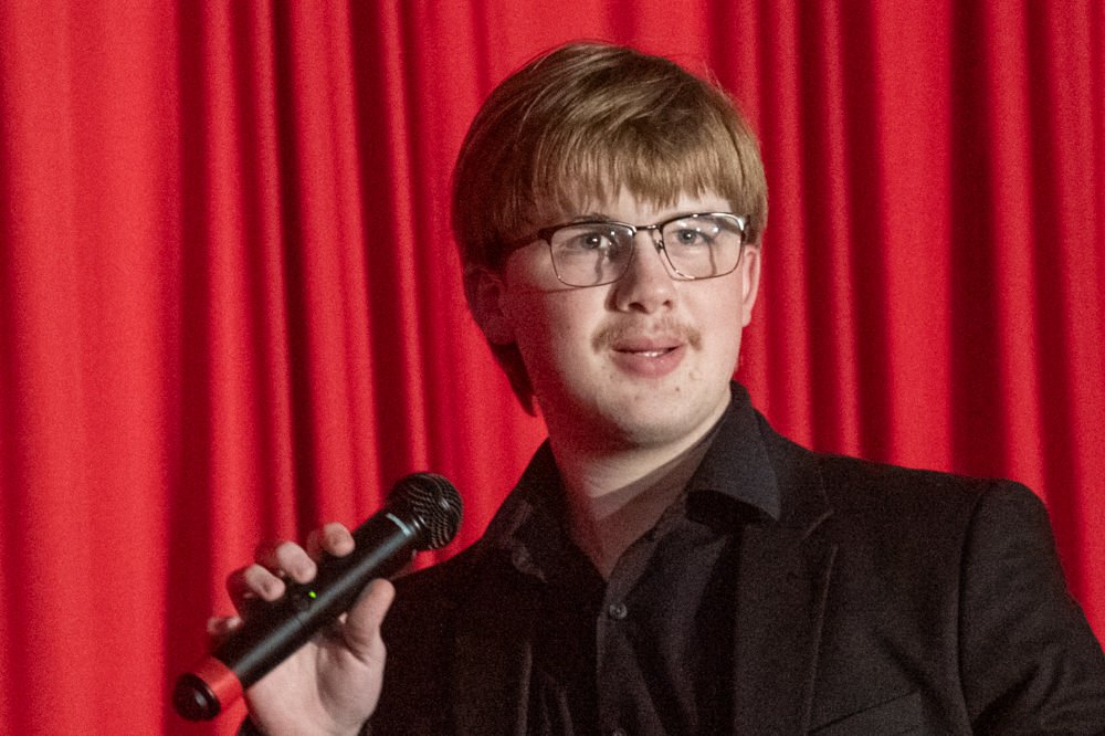 Young man with glasses speaking into a handheld microphone on a stage with a red curtain backdrop.