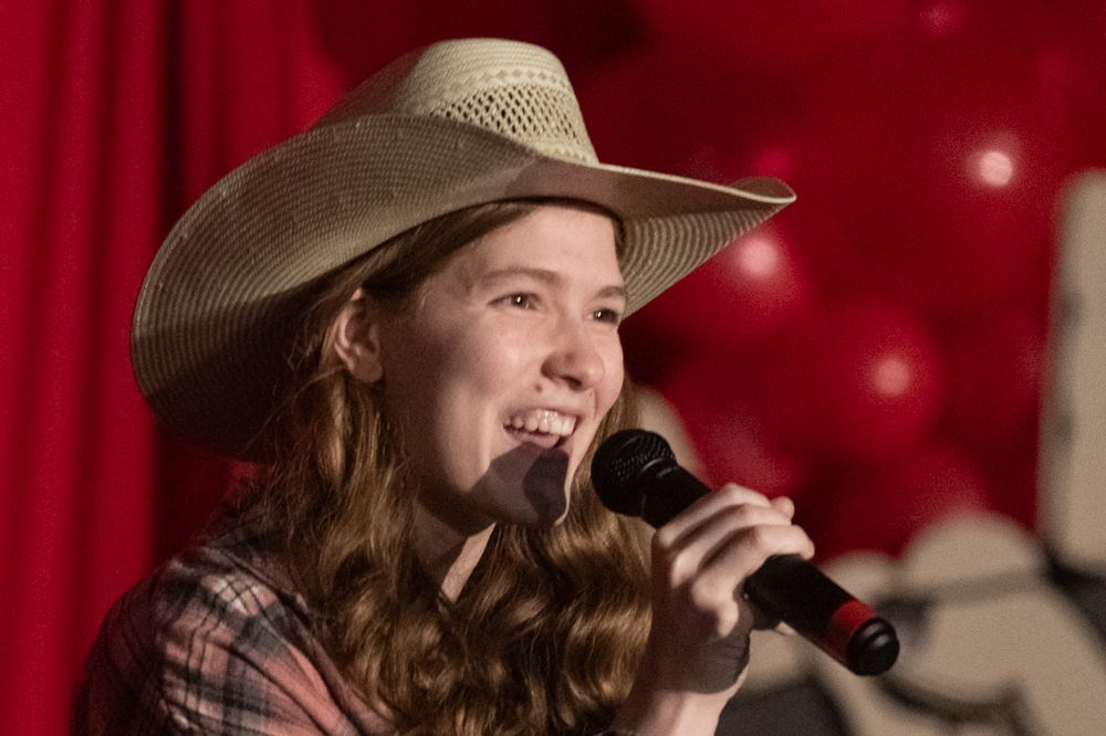 Young woman wearing a straw cowboy hat sings into a handheld microphone on a stage with a red backdrop and balloons behind her.