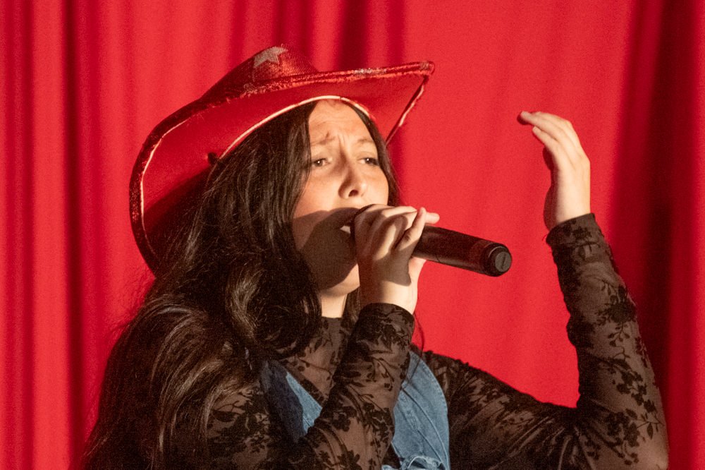Female singer performing on stage, wearing a red cowboy hat and blue overalls with black lace sleeves, singing into a microphone in front of a red curtain.