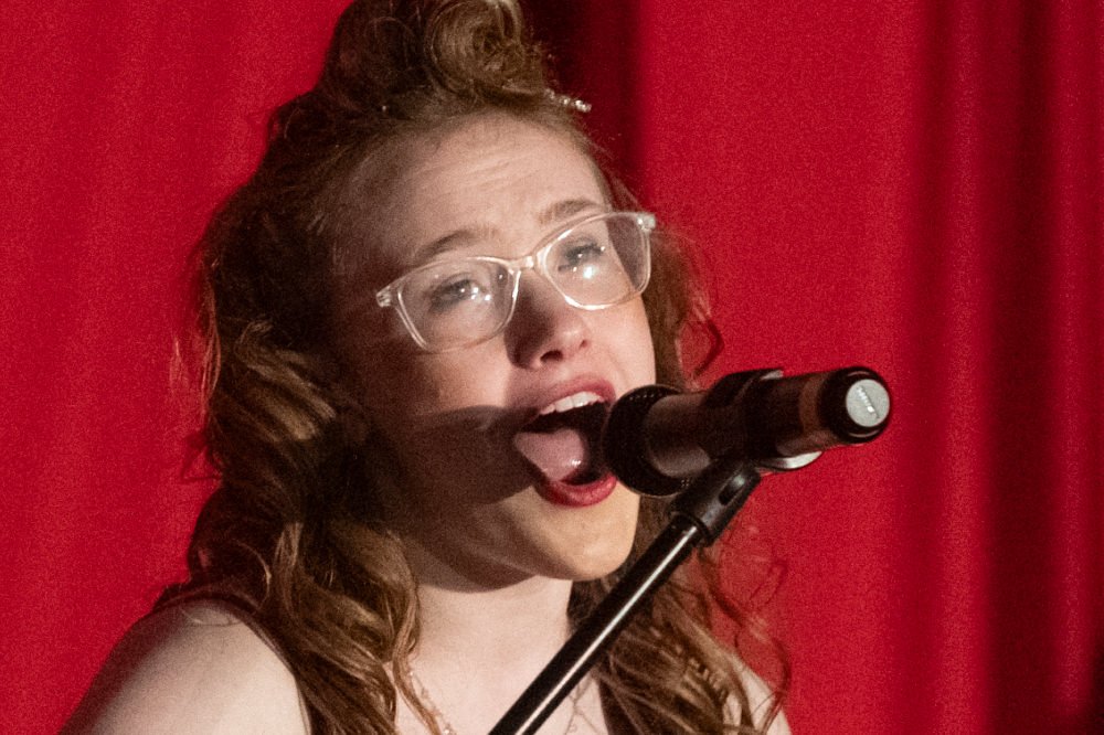 Young woman with clear glasses singing into a mounted microphone on a stage with red curtains in the background.