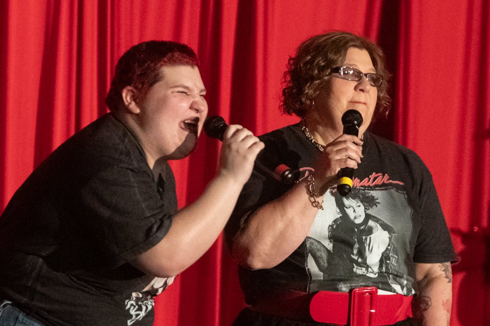 Two people perform on a stage in front of red curtains, each holding a microphone.