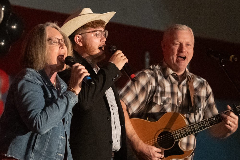 Three performers sing into microphones on a stage; a woman in a denim jacket, a man with a hat, and another man playing an acoustic guitar.