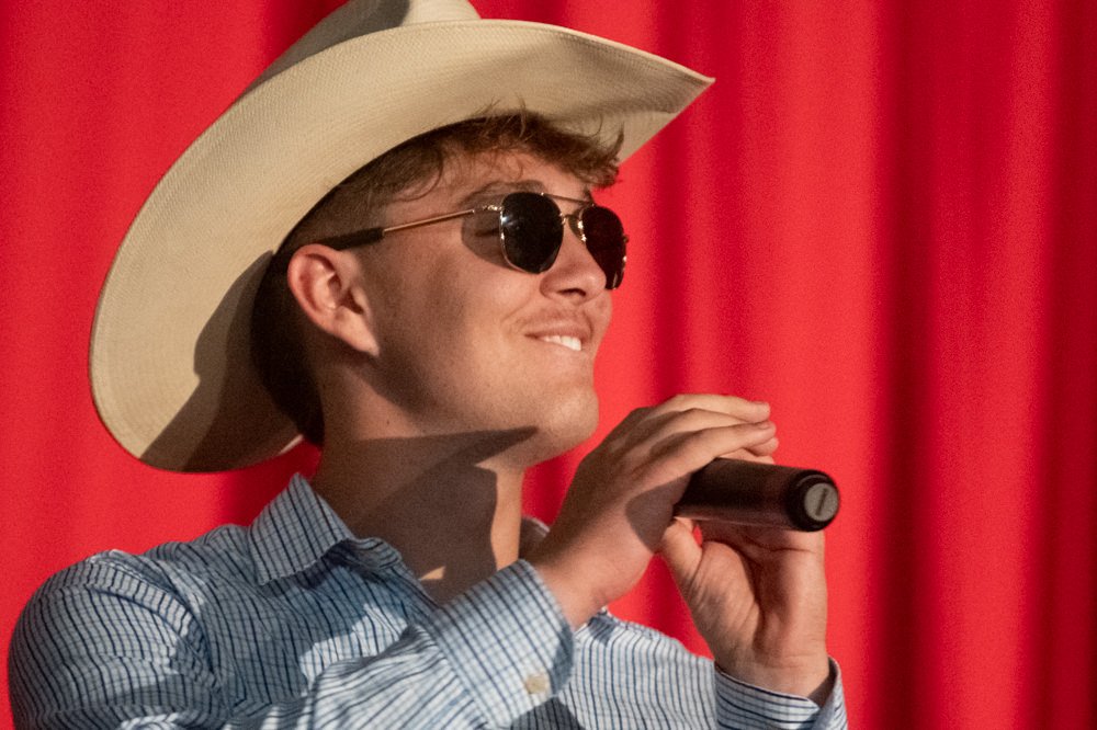 Male performer in a cowboy hat and sunglasses singing into a microphone on stage with a red curtain backdrop, wearing a blue checkered shirt.