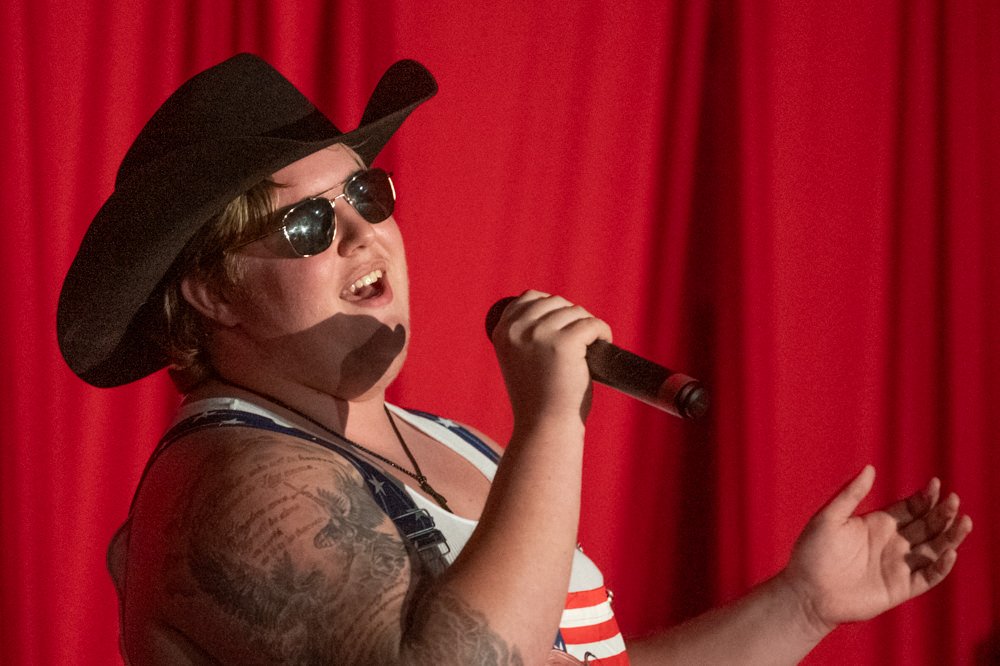 Singer wearing a black cowboy hat and sunglasses, holding a microphone on a red-curtained stage with tattoos visible on the arm.