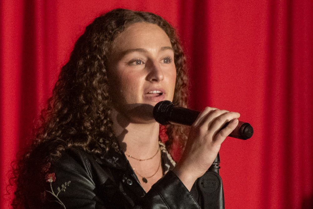 Young woman with curly hair singing into a microphone on a stage with red curtains behind her, wearing a black jacket with floral embroidery.