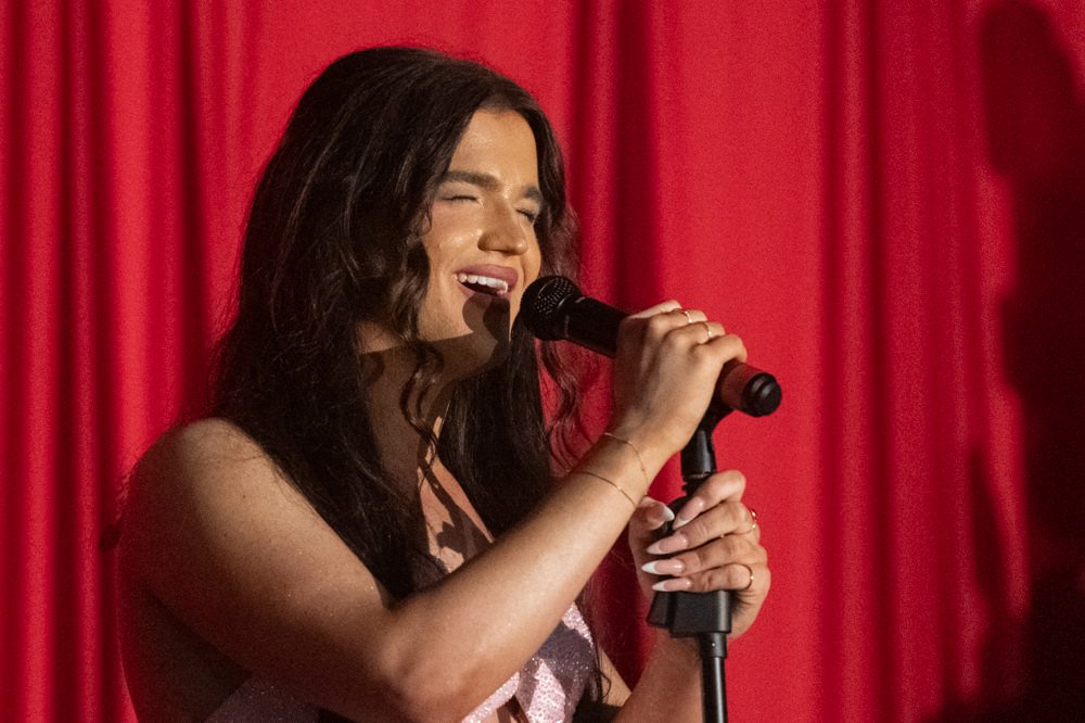 Female singer with long dark hair sings into a handheld microphone on stage, red curtain backdrop behind her, eyes closed.
