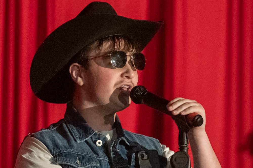 Young performer in a cowboy hat and sunglasses sings into a microphone on a red-curtained stage.