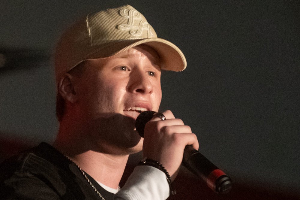 Man wearing a beige cap singing into a handheld microphone on stage.