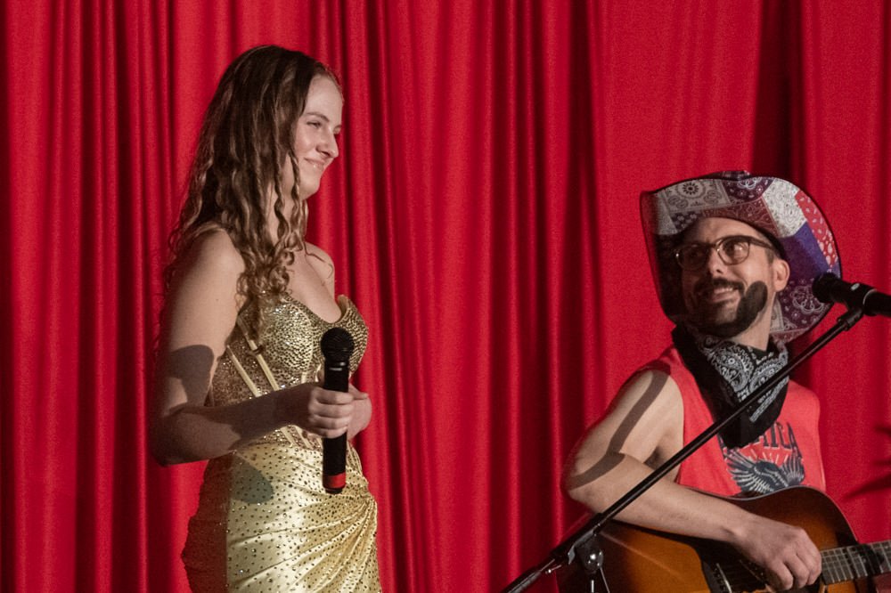 Woman in a gold sequined dress holding a microphone on stage with a red curtain backdrop, smiling at a guitarist beside her.