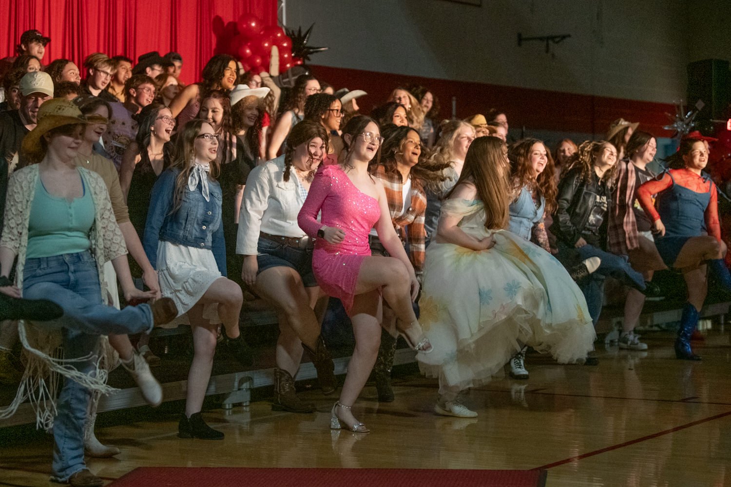 Group of students dancing on a school gym stage with red curtains and balloons in the background, wearing casual and festive outfits.