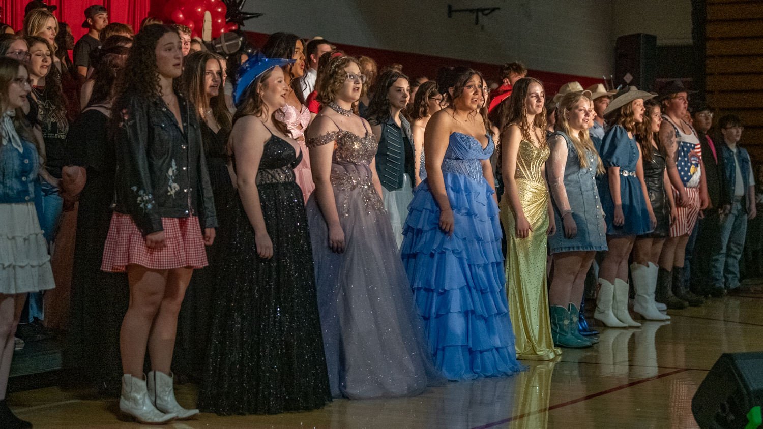 Line of students in formal dresses and outfits standing in a gymnasium on stage for a school event, facing forward.
