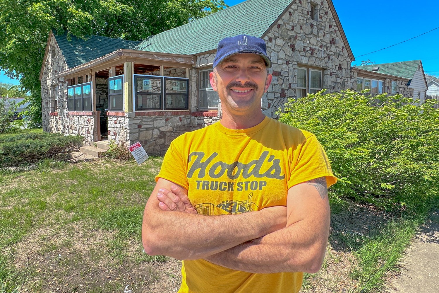 Smiling man in a bright yellow Hood's Truck Stop T-shirt stands with arms crossed in front of a stone storefront on a sunny day.
