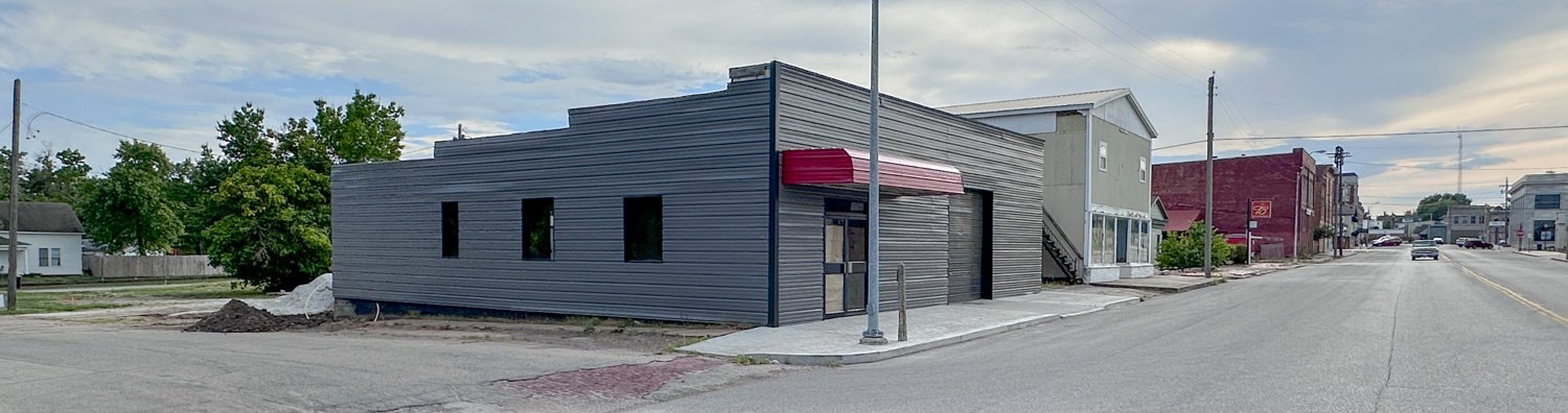 A gray, single-story building with a red awning on a quiet street, adjacent to older storefronts and a sidewalk under a cloudy sky.