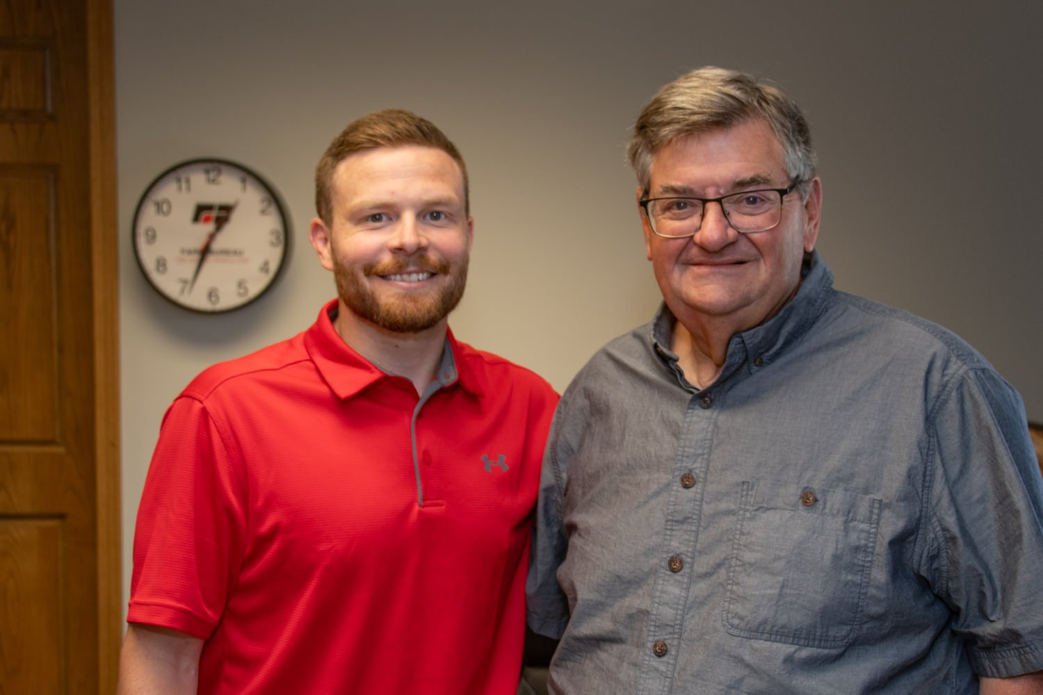 Two men pose together in an office: one in a red polo shirt and the other in a gray button-down, smiling at the camera with a clock on the wall behind them.