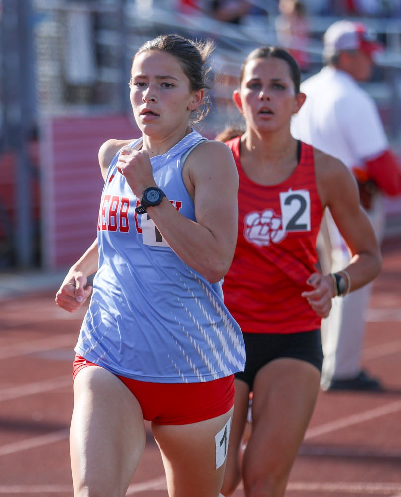 Two female runners race on a track, foreground runner in light blue top and red shorts, wearing a watch and bib 1.