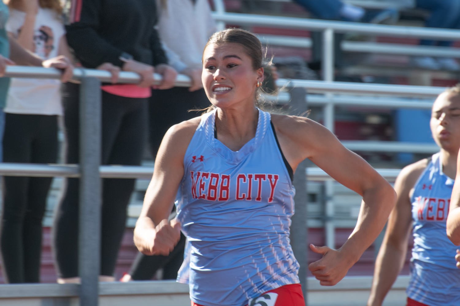 Female sprinter in a light blue WEBC CITY uniform runs on a track with spectators in the stands behind her.