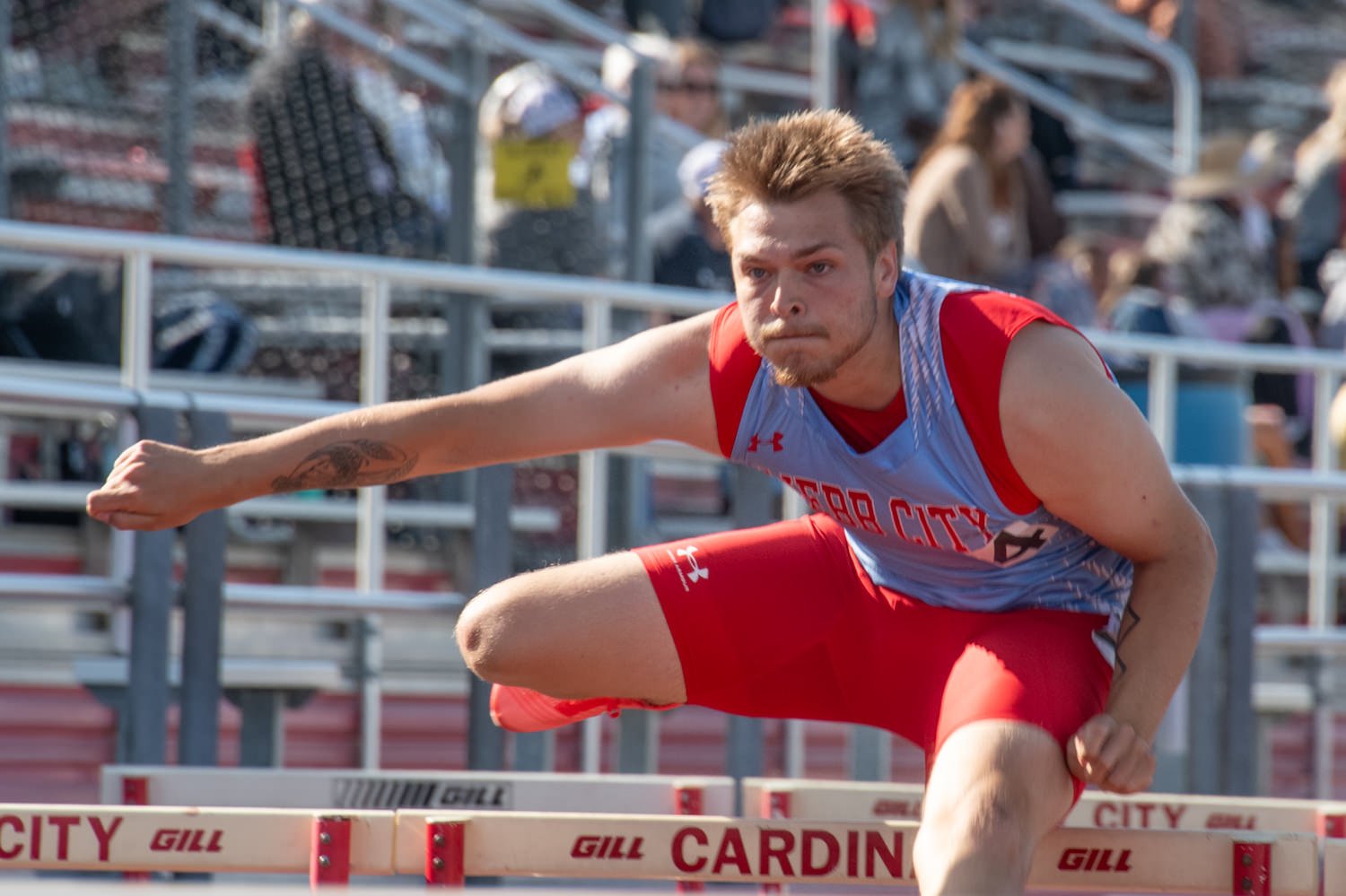 Male hurdler in a red-and-blue uniform leaps over a track hurdle, focused on the upcoming race.