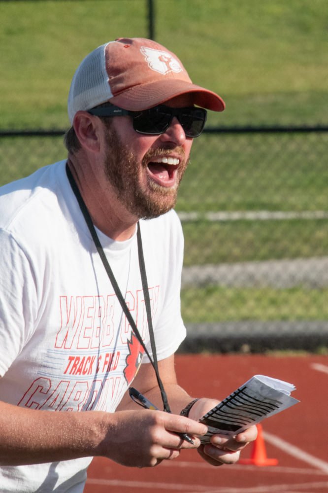 Coach on a track shouts instructions, wearing a pink cap, sunglasses, and a lanyard, holding papers and a pen clip.