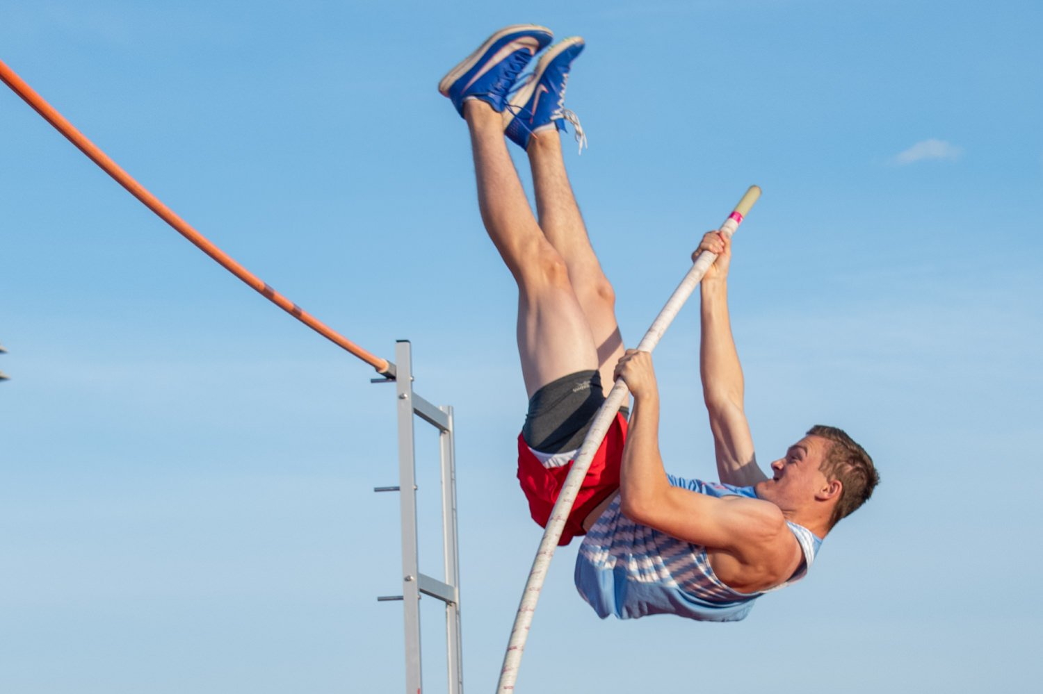 Male track and field athlete vaulting, upside down, using a long pole against a clear blue sky