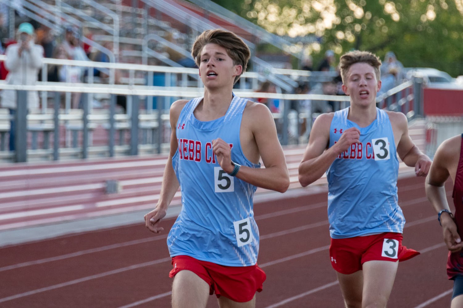Two male track runners in light blue jerseys and red shorts sprint on a red track, race numbers 5 and 3 visible, with spectators in the stands behind them.