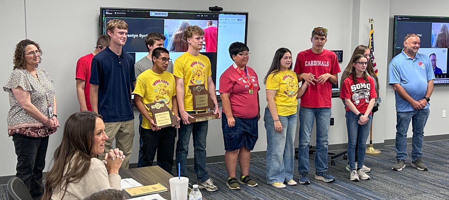Group of students standing in a row posing with trophies and medals after a competition, flanked by teachers in a classroom setting.