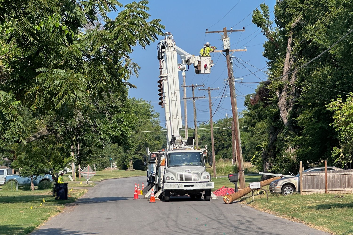 Utility worker in a bucket lift on a white service truck repairing electrical lines along a residential street; orange cones mark the work area.