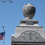 Stone monument with a carved urn on a stepped pedestal, blue sky behind and an American flag waving to the left.