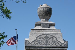Stone monument with a carved urn on a stepped pedestal, blue sky behind and an American flag waving to the left.