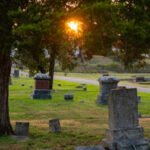 Sunset glow over a cemetery with weathered tombstones and tall trees framing the scene.