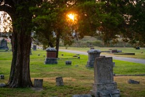 Sunset glow over a cemetery with weathered tombstones and tall trees framing the scene.