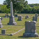 Cemetery with several tombstones and a tall obelisk, green grass and trees in the distance.