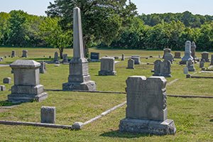 Cemetery with several tombstones and a tall obelisk, green grass and trees in the distance.