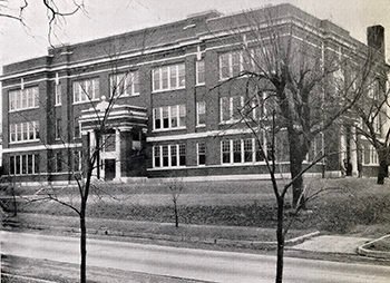 Two-story brick school building with a rounded central entrance, set back from the street, leafless trees in the foreground.