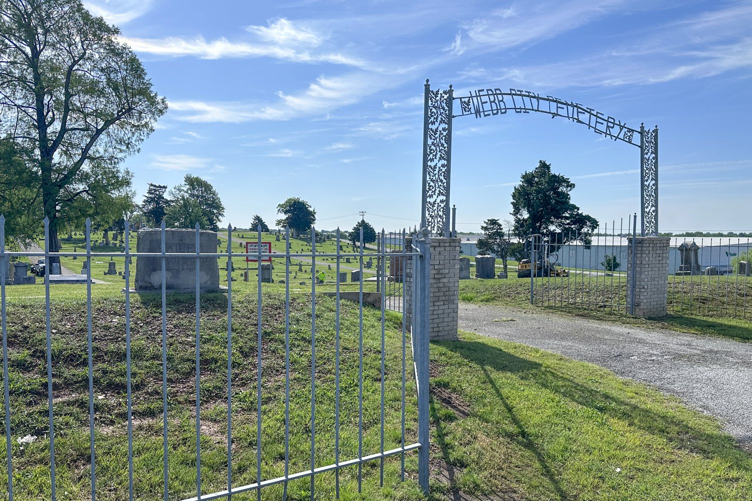 Decorative metal entrance arch labeled Webb City Cemetery with brick pillars, open gate leading to a grassy graveyard under a blue sky