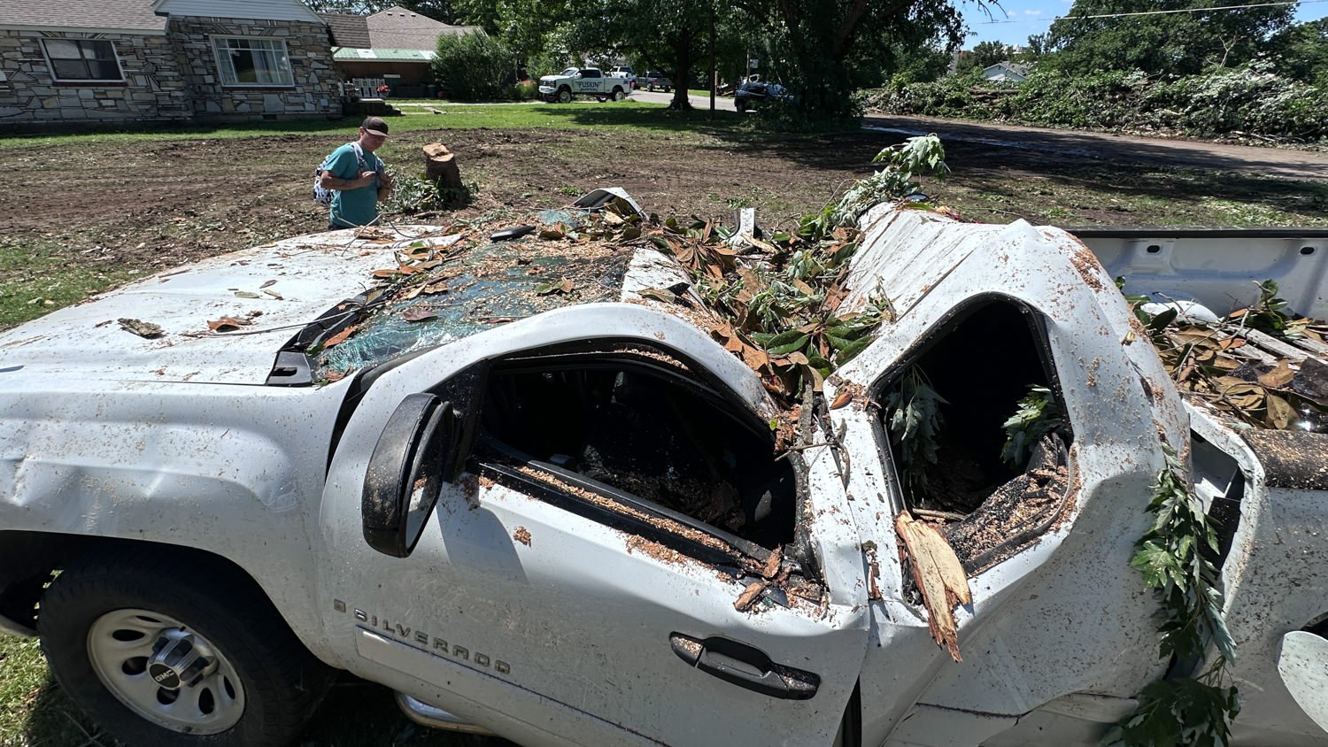 Damaged white Chevrolet Silverado with shattered windows and tree branches piled on the bed; man inspecting debris in a yard.