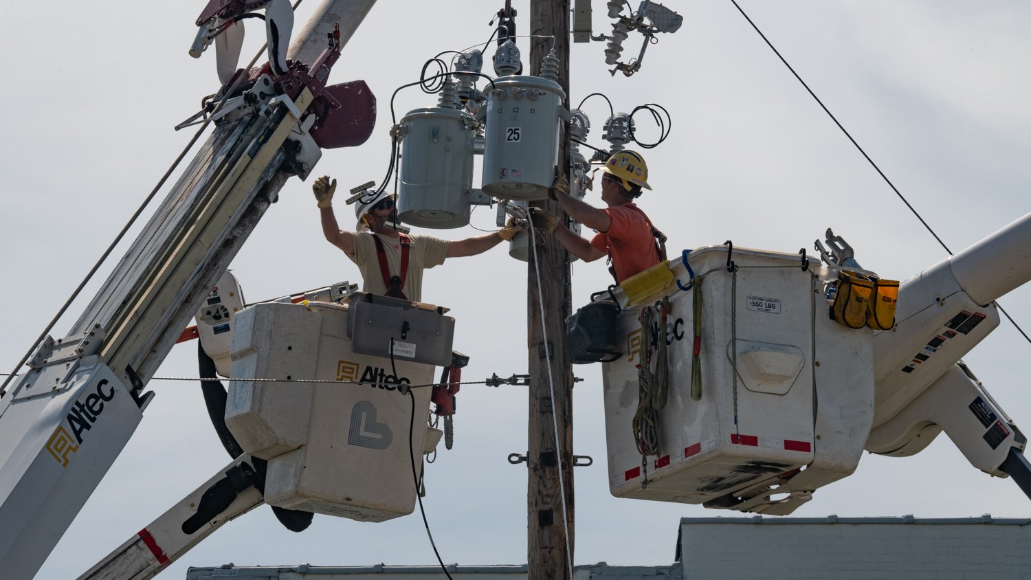 Two linemen in bucket trucks install or service electrical transformers on a utility pole with safety gear.