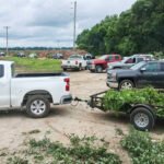 Pickup trucks parked in a gravel lot with a trailer loaded with green branches; a wooded area in the background under a cloudy sky.