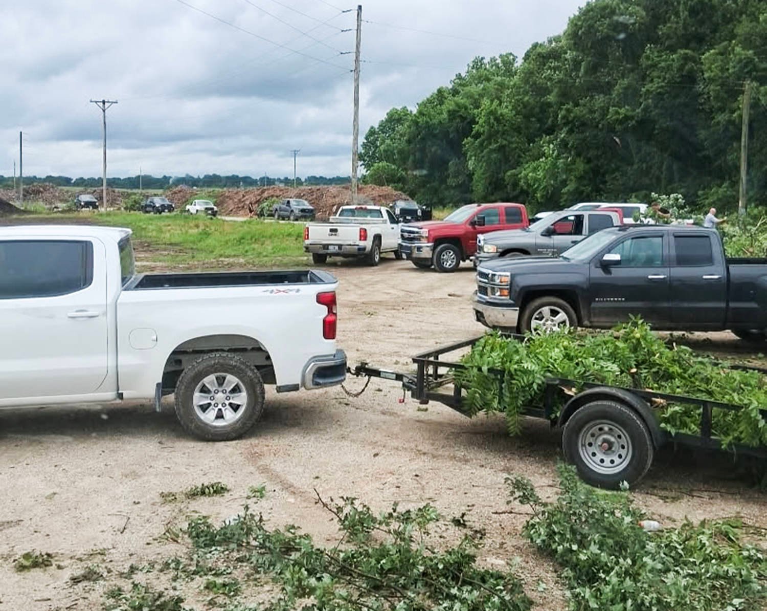 Pickup trucks parked in a gravel lot with a trailer loaded with green branches; a wooded area in the background under a cloudy sky.